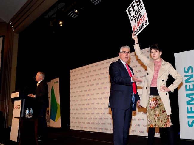 Protester Agnes Prest stand on the stage while the Prime Minister carries on.