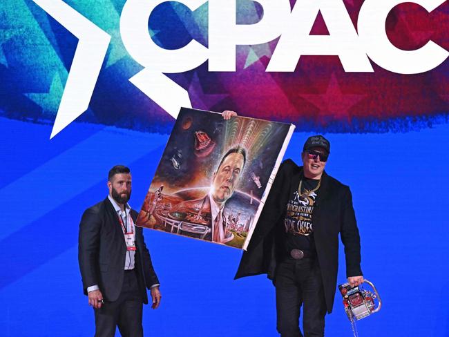 Elon Musk walks on stage holding a painting of himself and a chainsaw reading “Long live freedom, damn it” during the annual Conservative Political Action Conference in Maryland, Picture: AFP