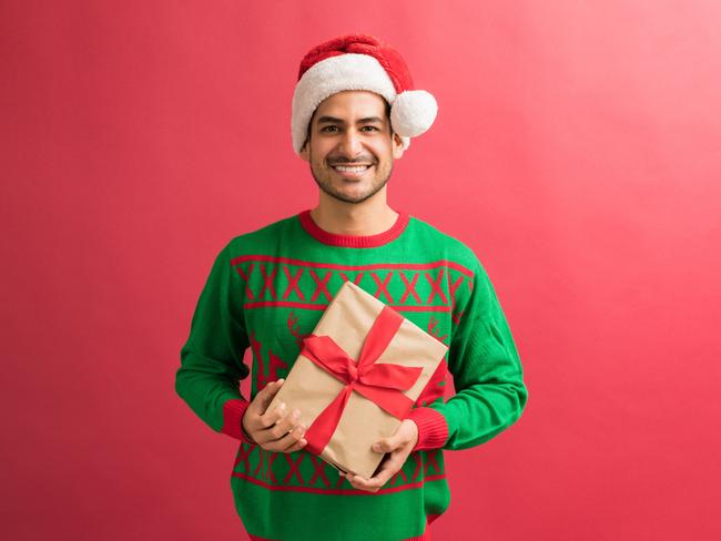 EMBARGO FOR TWAM 18 DEC 2021. FEES MAY APPLY. Full length of attractive young male holding Christmas gift while making eye contact against plain background