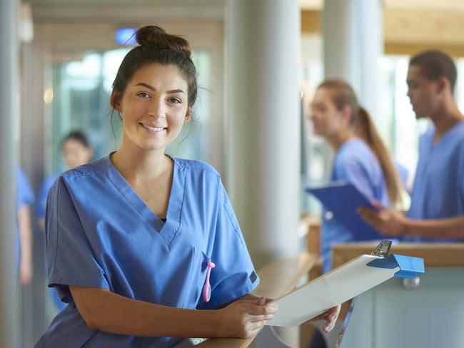 A young female nurse wearing blue scrubs and holding medical records is smiling to camera. She is standing on a busy corridor of a modern hospital . More male and female nurses are walking behind her .