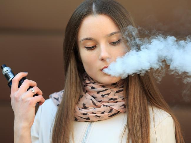 Vape teenager. Young pretty white girl in casual clothing smoking an electronic cigarette opposite modern brown background on the street in the spring. Bad habit. Vaping activity. Close up. i STOCK IMAGE