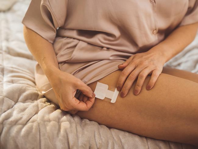 Mature woman waking up and using products for hormone replacement therapy. She is sitting on bed in pajamas and putting adhesive patch on leg. HRT