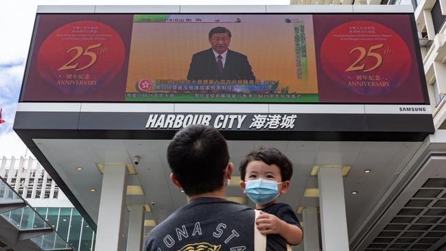 A man holding a child walks in front of a screen showing a live broadcast of Chinese President Xi Jinping speaking during a swearing-in ceremony for Hong Kong's new chief executive John Lee on July 1.