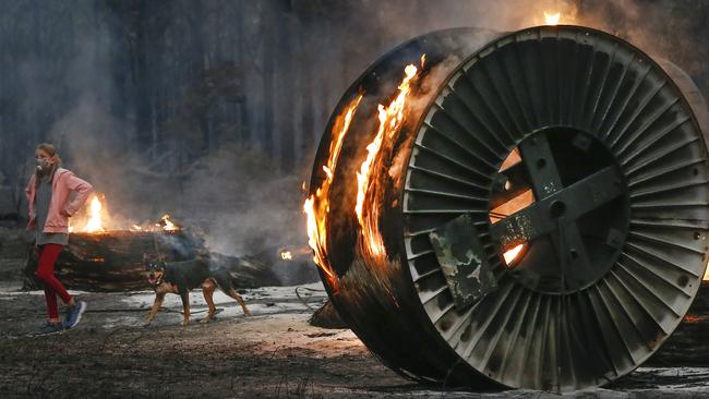 A young girl walking her dog past burning large cable spools along a scorched property in Mallacoota. Picture: David Caird