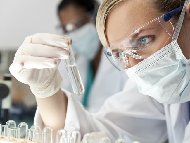 A blond medical or scientific researcher or doctor using looking at a clear solution in a laboratory with her Asian female colleague out of focus behind her.