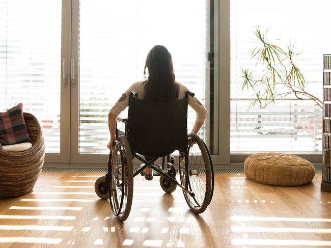 Beautiful young disabled woman in wheelchair at the window at home in her living room. Rear view.