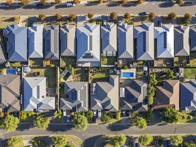 Aerial view directly above new outer suburban/semi-rural housing development with single-level housing between two streets with t-intersections and orange coloured street trees.  Mostly gray metal roofing, landscaped front and back yards, some solar panels, cars and motor vehicles parked in driveways and street, some street and parkland trees, water tanks in backyards, sheds and garages, swimming pools.  Mount Barker, South Australia; property investment housing money generic