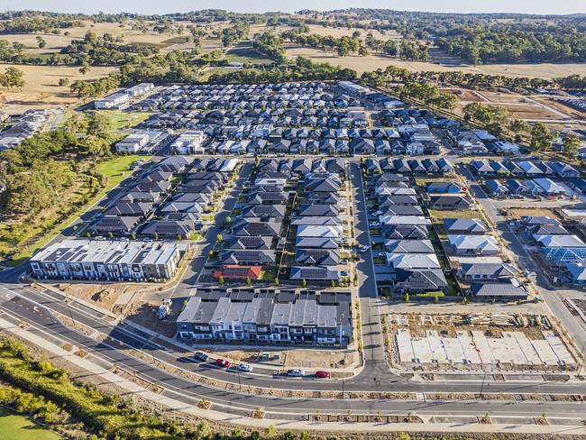 Standing out from the crowd: one red roof amongst rows of houses of various architectural styles with shades of grey in rural urban sprawl. Houses under construction alongside newly-completed buildings in rural setting.  Tightly-packed houses with small front and back yards and few if any trees. New divided arterial access road under construction in foreground, railway line in background. Mount Barker, South Australia; property investment Australian housing money generic