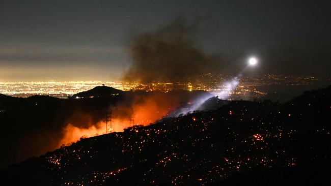 The light of a fire fighting helicopter illuminates a smouldering hillside as the Palisades fire grows near the Mandeville Canyon neighbourhood and Encino, California. Picture: Patrick T. Fallon / AFP