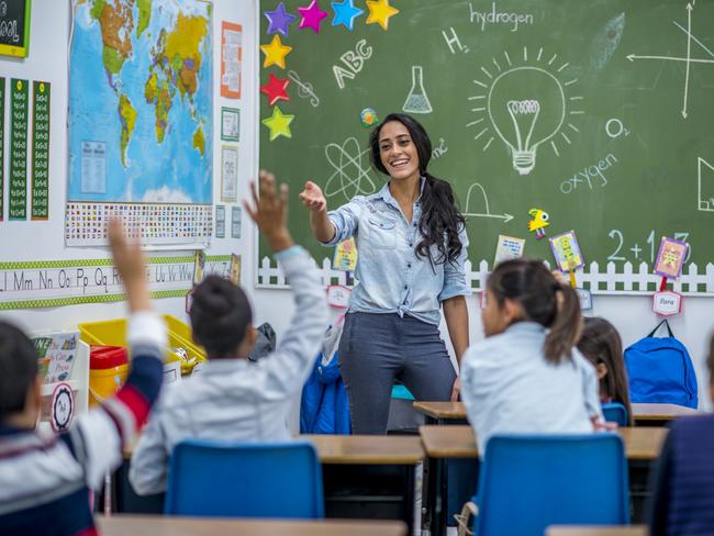 An Ethnic teacher is leading a class of elementary school children. There are various posters on the wall, and drawings on the chalkboard. Students are putting up their hands to answer a question.Picture: iStock