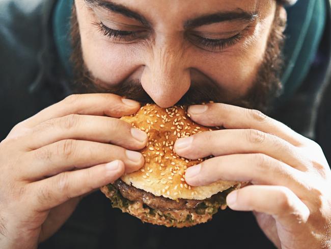 Closeup front view of a mid 20's man biting on a juicy burger outdoors. He has brown beard and mustache. He's wearing winter jacket and a cap.