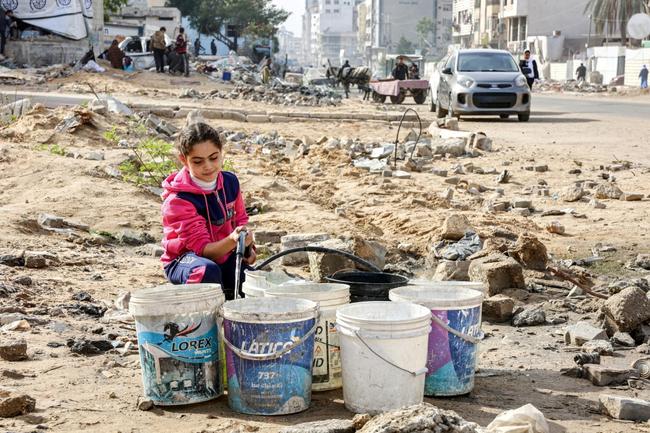 A girl fills plastic buckets with water from a hose in an empty area littered with debris in Gaza City