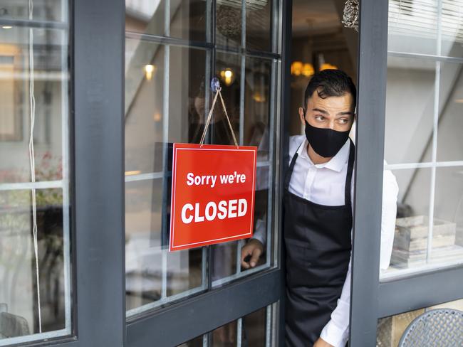 Male restaurant employee wearing protective face mask, watching behind the half-open door where is hanging a signboard "sorry, we're closed".