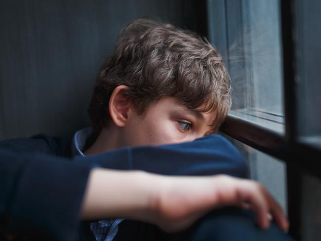 Pensive sad boy teenager with blue eyes in a blue shirt and jeans sitting at the window and closes his face with his hands.