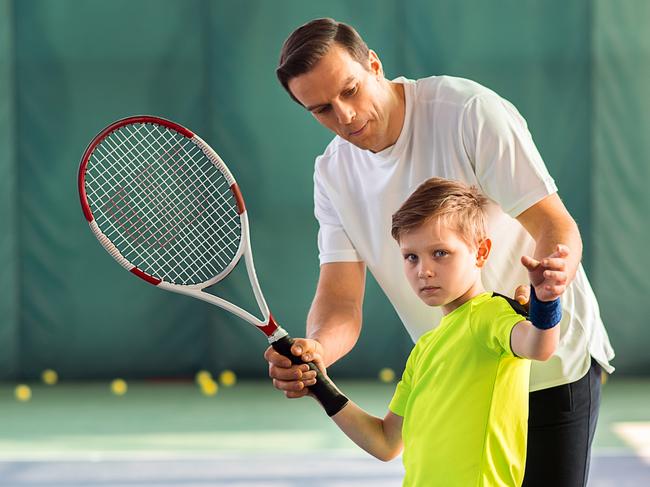 Professional male tennis player is explaining to child how to play tennis. He is adjusting racket in childish hands. Boy is standing and looking forward with seriousness. Portrait