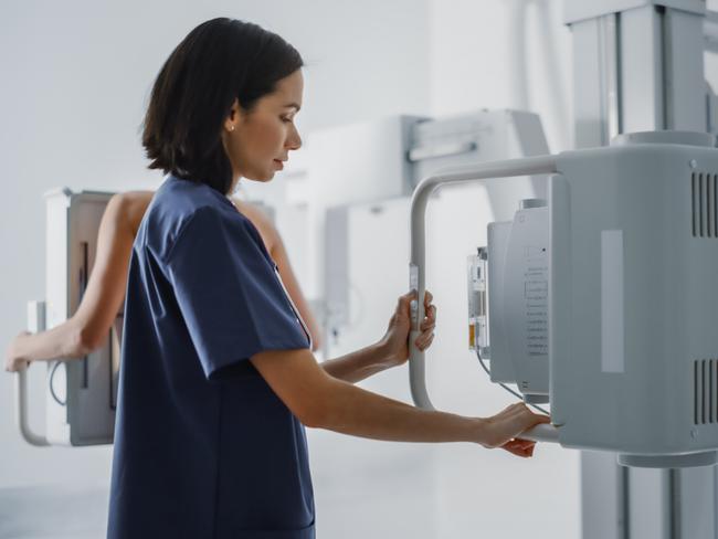 Hospital Radiology Room: Beautiful Multiethnic Woman Standing Topless Next to X-Ray Machine while Female Latin Nurse Adjusts it. Healthy Patient Undergoes Medical Exam Scanning with the Doctor's Help.