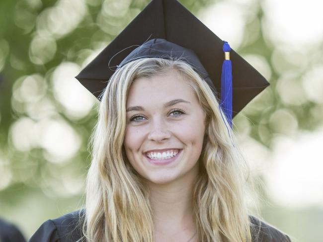 A multi-ethnic group of university students are outdoors at a graduation ceremony. They are wearing graduation robes and caps. They are walking away from the camera, and a Caucasian woman is turned to smile at the camera while holding her diploma.