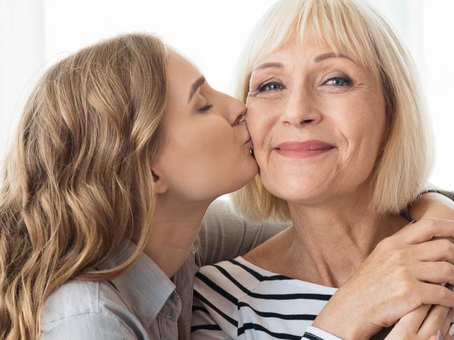 Young daughter kissing senior mother on the cheek, standing against window at home, copy space