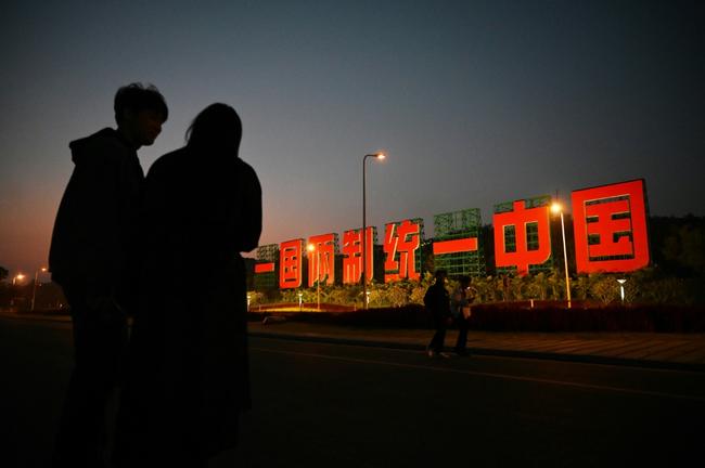 A couple stands near a giant propaganda slogan in China's Xinmen which reads 'One Country, Two Systems, Unify China', visible from Taiwan’s Kinmen Island