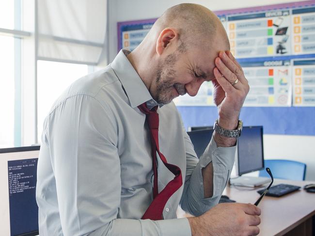 A teacher sits on the edge of a desk in an empty classroom, he is stressed