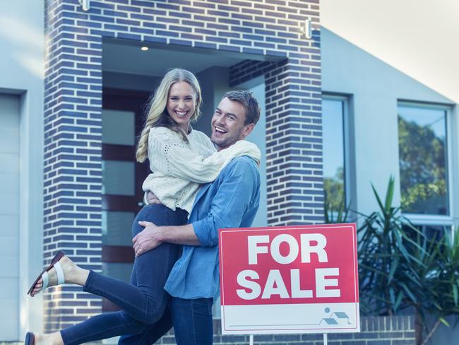 Couple in front of a new home. They are standing next to a for sale sign. They are both wearing casual clothes and embracing. They are smiling and he has a beard. He is lifting his wife off the ground with joy. The house is contemporary with a brick facade. The front door is also visible. Copy space