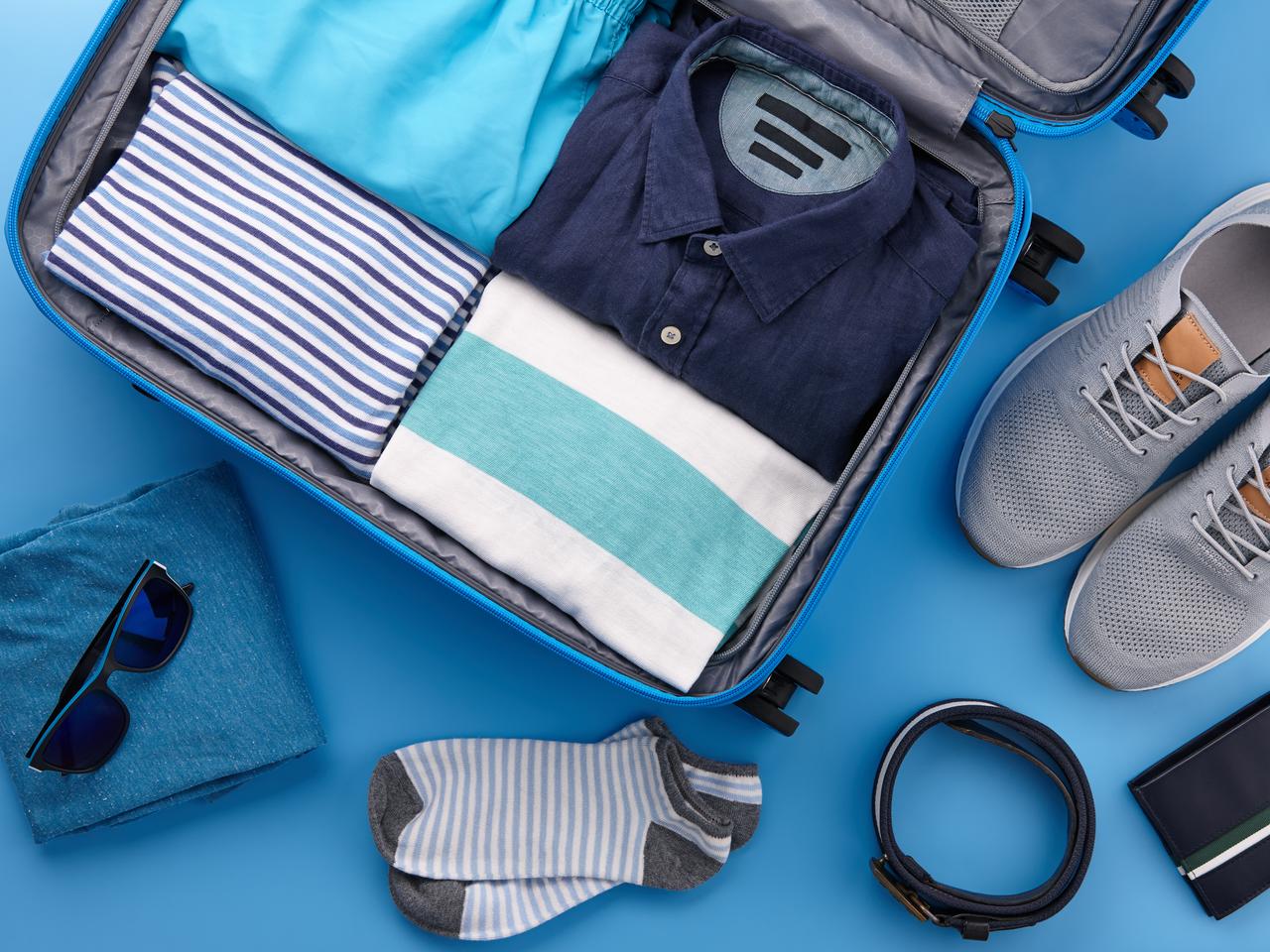 Packing men's summer clothes and accessories in blue travel suitcase on blue background, top view