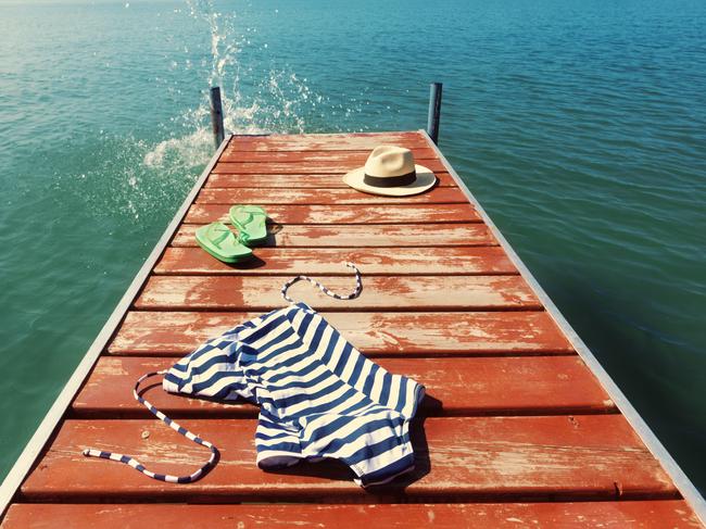 Skinny dipping and jumping in the lake concept, suggested by blue and white striped woman’s swimsuit, flip-flops, and Panama hat scattered on an old wooden boat pier. Water spray splashes up at the end of the dock, as if a naked person went diving into fresh water for a swim.