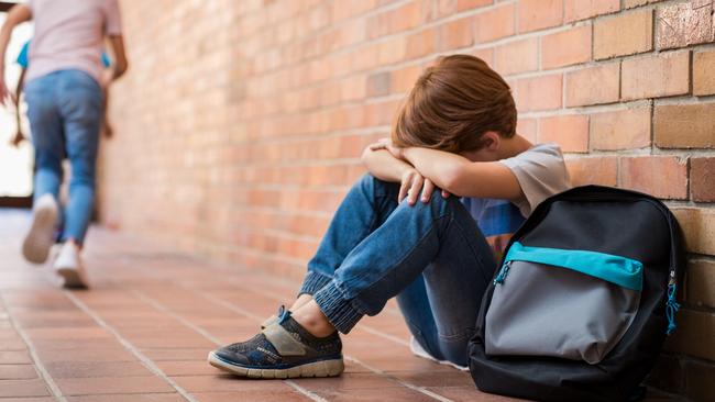 Little boy sitting alone on floor after suffering an act of bullying while children run in the background. Sad young schoolboy sitting on corridor with hands on knees and head between his legs.