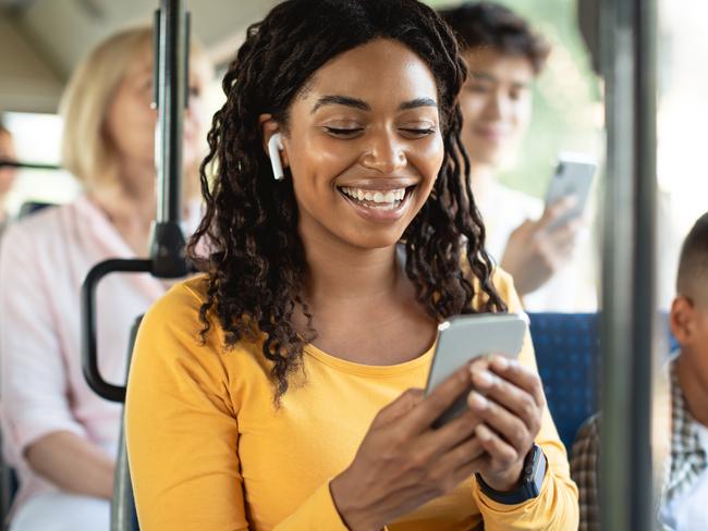 Laughing black lady using mobile phone sitting on city bus seat, happy passenger in wireless headphones listening to music, watching video or movie, making online call, enjoying travel ride