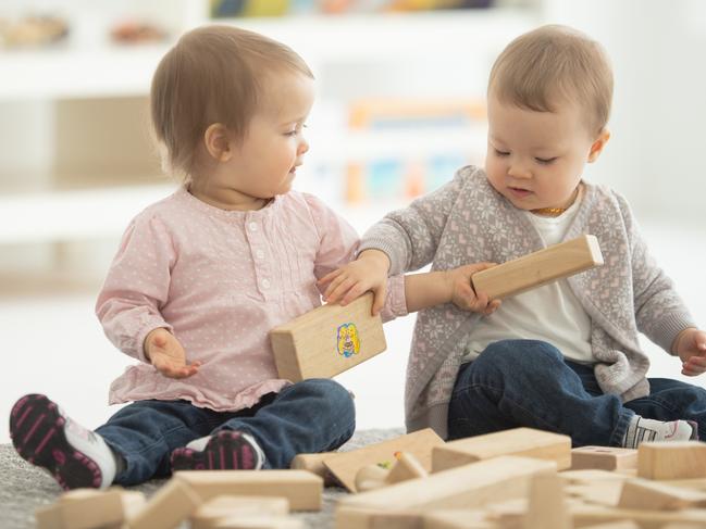 Two baby caucasian girls play with wooden blocks on a rug.