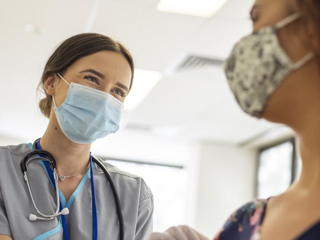 Shot focused on a kind looking nurse wearing scrubs, a stethoscope, white rubber gloves and a protective face mask. She is smiling at the nervous patient with her eyes. The nurse is prepping the patients arm before she injects her with the COVID-19 vaccine.