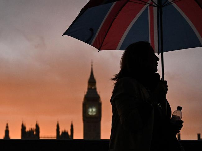 A member of the public holding an a Union flag themed umbrella stands in the queue, with the Palace of Westminster, house of Parliaments and Elizabeth Tower, commonly referred to as Big ben, on the background at sunset, as she waits in line to pay her respects to the late Queen Elizabeth II, in London on September 18, 2022. - Britain was gearing up Sunday for the momentous state funeral of Queen Elizabeth II as King Charles III prepared to host world leaders and as mourners queued for the final 24 hours left to view her coffin, lying in state in Westminster Hall at the Palace of Westminster. (Photo by Marco BERTORELLO / AFP)