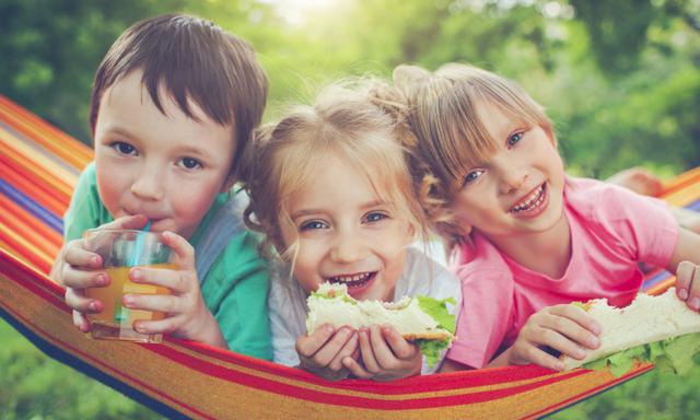 Three happy children eating sandwiches and drinking lemonade in hammock in a park or back yard in summer