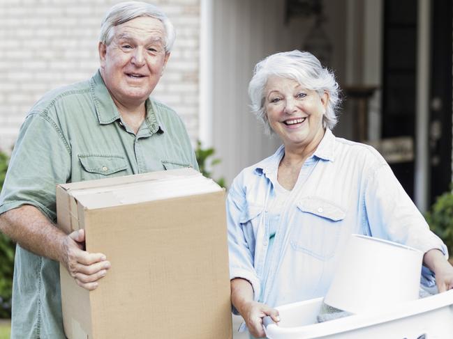 A senior couple relocating, downsizing to a smaller home. They are carrying a box and laundry basket into their new house, looking at the camera and smiling. They are in their 60s.