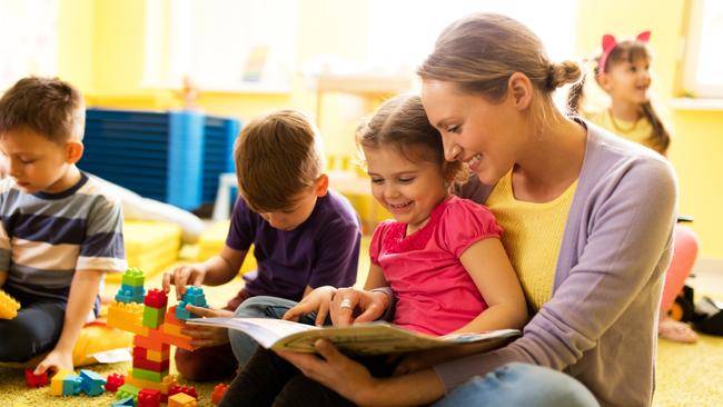 Happy little girl sitting in teacher's lap at preschool and reading a book while other children are playing.