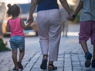 Rear photo of mother holding hands of son and daughter while walking on the street. Selective focus on models. Shot during summer time in outdoor with a full frame mirrorless camera.