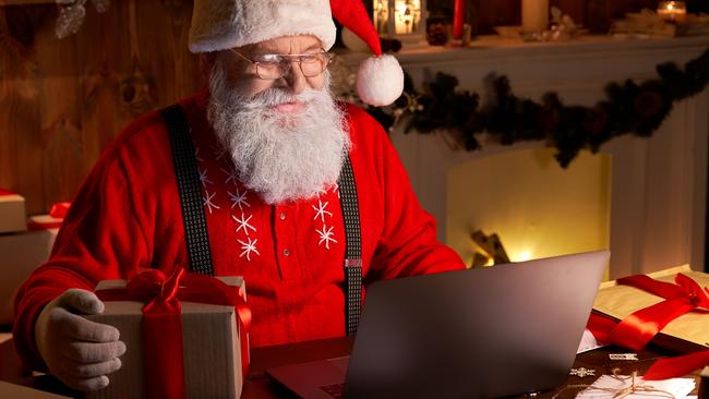 Happy old Santa Claus wearing hat holding gift box using laptop computer sitting at workshop home table late on Merry Christmas eve. Ecommerce website xmas time holiday online shopping e commerce sale