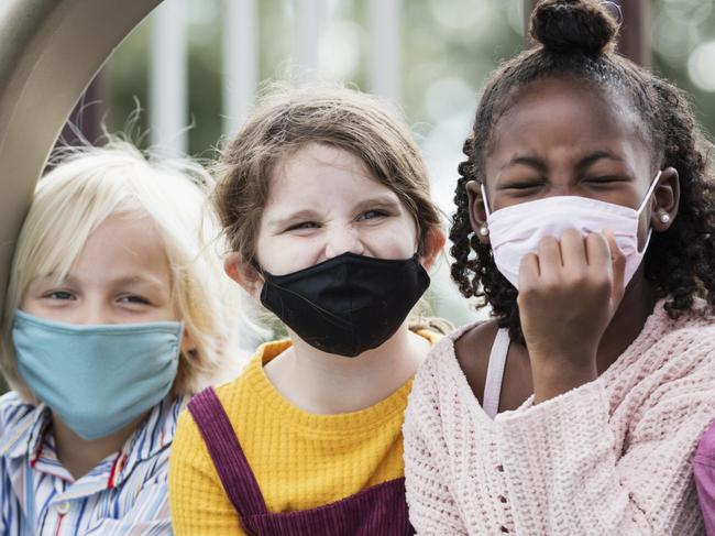 A multi-ethnic group of four children, 5 to 7 years old, playing together on a playground during school recess. They are sitting side by side on two slides, looking at the camera. They are all wearing masks, back to school during the COVID-19 pandemic, trying to prevent the spread of coronavirus.