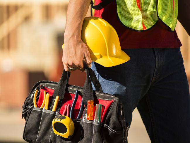 A construction worker busy working at a job site. He holds a tool box full of tools and a hard hat. Framed house, building in background. He is wearing a safety vest.