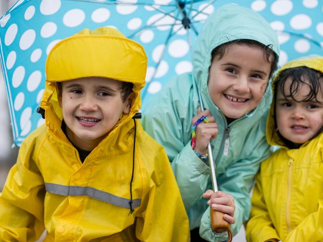 Three Ethnic siblings are outdoors during a rain shower. They are wearing rain jackets with hoods. They are sheltering under a blue spotted umbrella.