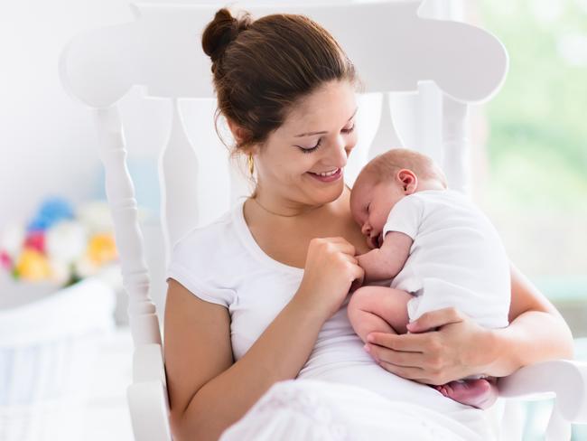 Young mother holding her newborn child. Mom nursing baby. Woman and new born boy relax in a white bedroom with rocking chair and blue crib. Nursery interior. Mother breast feeding baby. Family at home