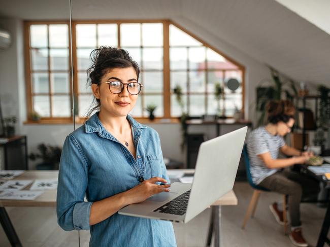 Portrait of young smiling woman in creative office, standing, holding laptop, looking at camera.