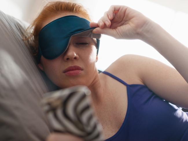 Young woman with sleep mask waking up in bed and snoozing alarm clock on cell phone, mobile telephone. Girl sleeping in bedroom at home and waking up after receiving text message or call on smartphone