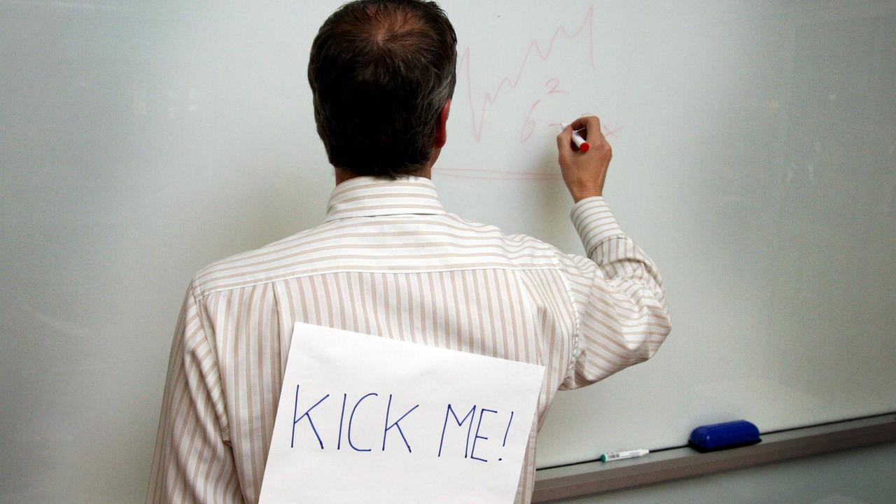 Man writing on a white board with a "kick me" sign taped to his back.