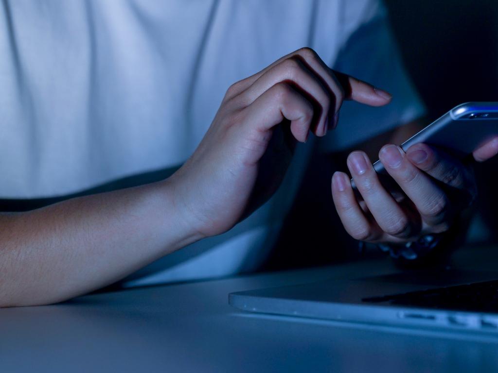 close up young man hand holding smartphone for access by unlock on screen and using laptop for register website on internet in the late night , internet addiction concept