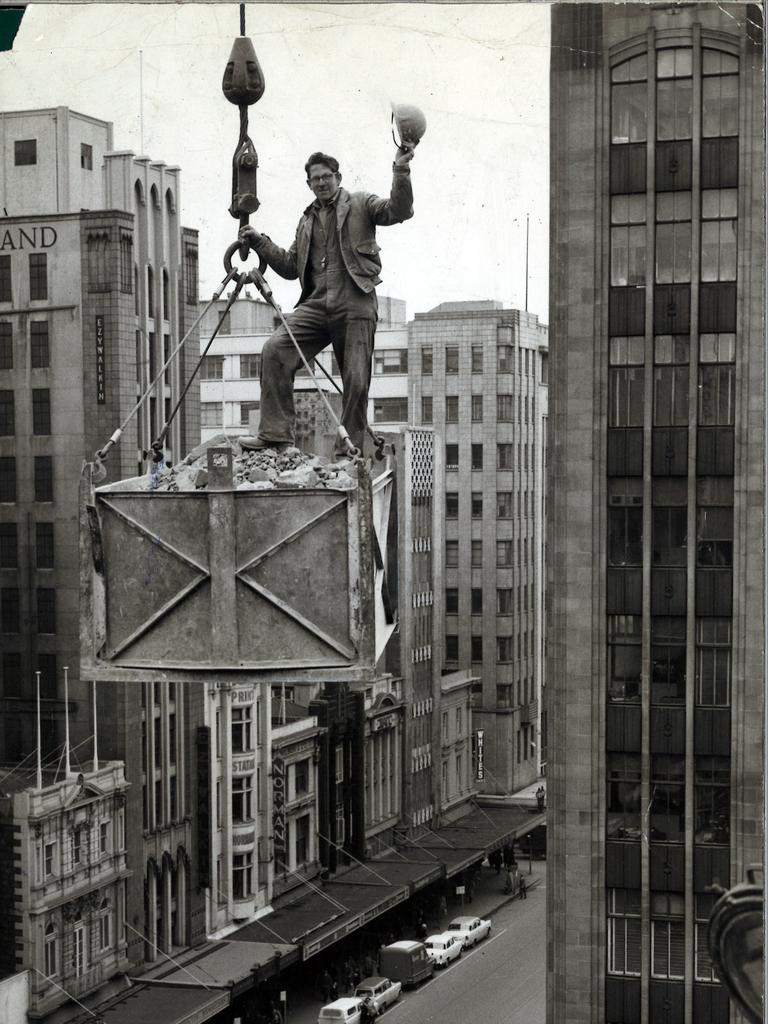 1960. Bob Schutt, 20, of Footscray, waves his tin helmet merrily as a crane pulls him and his load skyward at the CML Building above Elizabeth St, Melbourne.