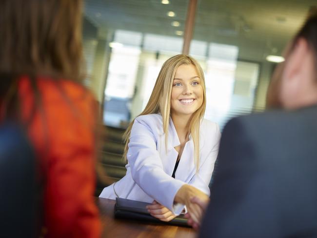 a young graduate sits across the table from her interview panel  full of confidence and positivity energy . She is holding her cv and smiling at the interview panel before her and shakes hands with the new boss.She is wearing blue trousers with an orange suit jacket . in the foreground we can see the back of two of the panel , a man and a woman.
