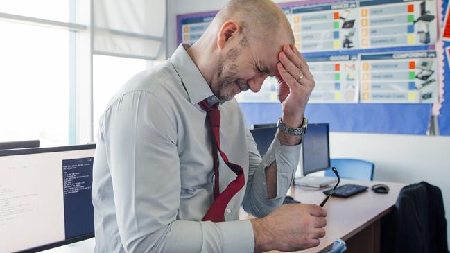 A teacher sits on the edge of a desk in an empty classroom, he is stressed