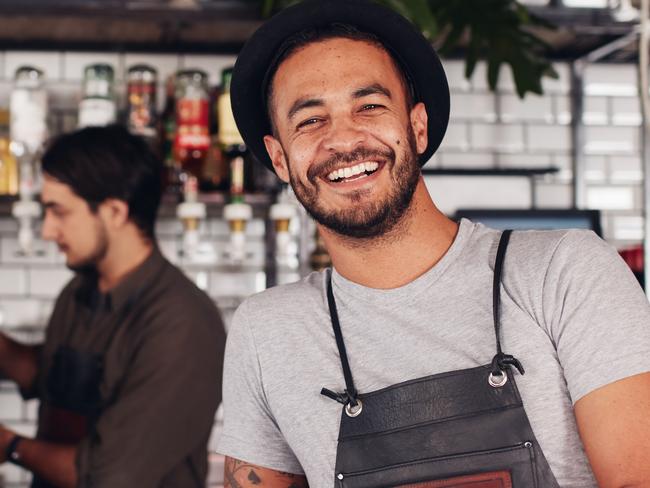 Coffee shop worker smiling to camera, standing at the counter. Happy young man in apron and hat leaning to cafe counter, with waiter working in background. For Safety First feature