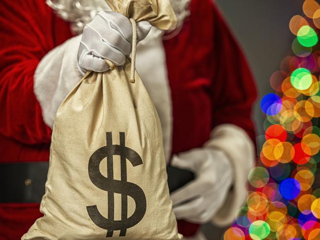 Santa Claus holding a large canvas money bag with black dollar sign symbol and in the background the bokeh of multi-colored Christmas tree lights